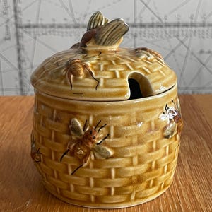 May include: A ceramic honey pot with a lid, featuring a woven basket design and several detailed bee embellishments. The pot is a warm, golden-brown colour, with a small opening for a spoon. The lid has a bee-shaped handle.