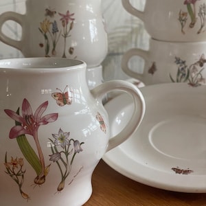 May include: White ceramic mugs and a saucer featuring floral and butterfly illustrations. The mugs have large handles and are stacked. The floral designs include pink, yellow, and blue flowers. The saucer has a butterfly design.