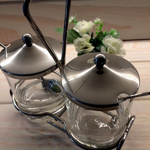 May include: A stainless steel condiment set with two clear glass jars, each with a lid and a small spoon. The set is held in a metal frame with a handle. A small bouquet of white flowers is in the background.