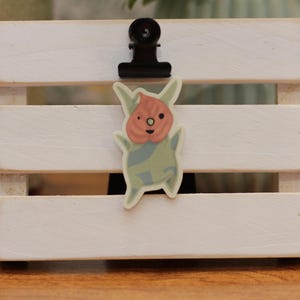 May include: A sticker of a cartoon creature with a green and blue body and a pink head. The creature has two large eyes and a small mouth. The sticker is on a white wooden surface.