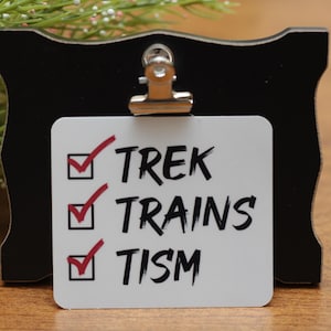 May include: A black wooden frame with a white checklist card clipped to it. The card has the words "TREK", "TRAINS", and "TISM" with red check marks next to each word. The background is a wooden surface with greenery.