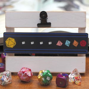 May include: A white wooden crate with a black clip holding a black sign with colorful geometric shapes. Various polyhedral dice in different colors and designs are scattered in front of the crate.