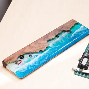 May include: A blue and white resin keyboard wrist rest with a wood border. The resin features a wave design with white foam and small red and white boats.