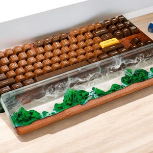 May include: A wooden keyboard wrist rest with a clear resin top featuring a green and white landscape design. The keyboard is brown with yellow keycaps and a white "SPACE BAR" key.