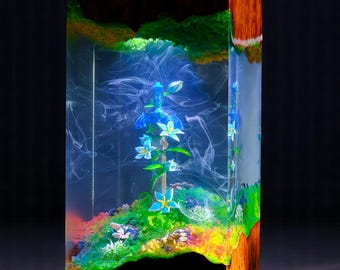 Puede incluir: Una pieza de arte rectangular de resina con marco de madera. En el interior, un vibrante paisaje en miniatura presenta flores azules, musgo verde y acentos coloridos. La resina transparente permite el paso de la luz, creando un efecto brillante.