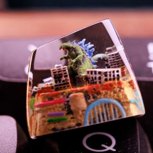 May include: A clear resin keycap with a miniature Godzilla destroying a city. The keycap is on a black keyboard with a "Q" key visible.