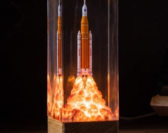 May include: A rectangular resin lamp featuring a miniature orange and white NASA rocket model encased inside. The base of the rocket is surrounded by a glowing, textured orange light effect resembling a launch plume, set on a wooden block base.