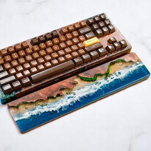 May include: A wooden keyboard with brown and beige keys and a matching wooden wrist rest featuring a blue and green resin design with white waves.