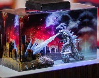 May include: A rectangular resin diorama featuring a Godzilla figure battling a city. The scene is set against a backdrop of a fiery sky and city buildings. The diorama has a black wooden top and is illuminated with a blue light.