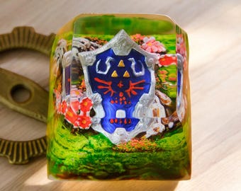 Puede incluir: Un keycap de resina transparente con un diseño de paisaje verde, marrón y blanco. El keycap presenta un escudo plateado y azul con un diseño rojo y dorado. El escudo está rodeado de flores y un fondo verde y musgoso.