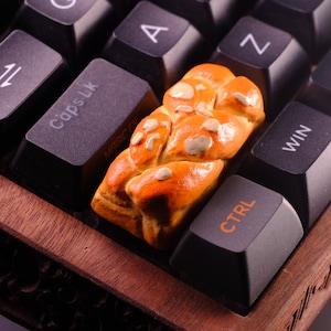 May include: A close-up of a keyboard with a braided bread-shaped keycap. The keycap is orange and brown with white sprinkles. The keyboard is made of wood and has black keys with white lettering. The keys are labeled with letters, numbers, and symbols. The keycap is on the CTRL key.