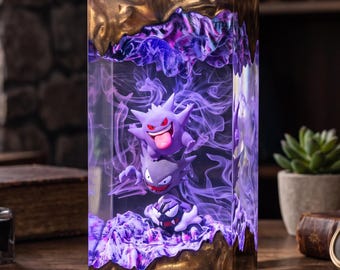 Puede incluir: Una lámpara rectangular de resina con tres figuras Gengar moradas apiladas dentro de material transparente. Las secciones superior e inferior tienen un acabado de bronce metálico texturizado. El interior brilla con una luz morada vibrante.