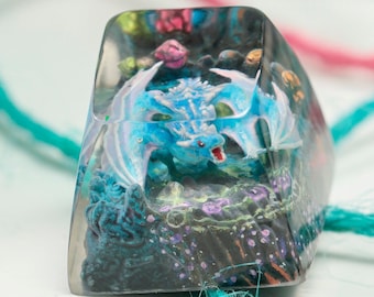 May include: A clear resin keycap with a blue dragon figurine inside. The dragon is surrounded by a dark blue and purple background with white and pink accents.