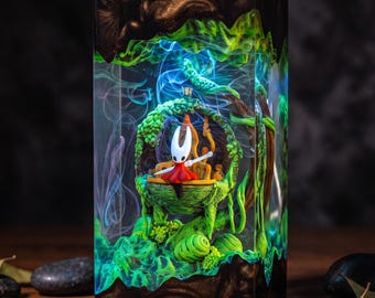 Puede incluir: Una lámpara rectangular de resina y madera que presenta una escena en miniatura con un personaje blanco y rojo sentado sobre una estructura verde cubierta de enredaderas. El interior brilla con luz verde y efectos de humo azul, enmarcado en madera oscura.