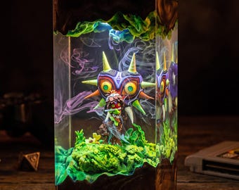 May include: A rectangular resin lamp featuring a miniature Majora's Mask figure suspended inside. The interior displays a green mossy base, swirling purple and white smoke effects, and internal LED lighting that illuminates the mask's vibrant colors.