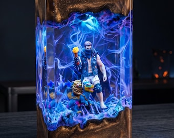 May include: A rectangular resin lamp featuring a dark bronze-colored top and bottom frame. The center contains a clear, glowing blue and purple swirling liquid effect surrounding a miniature figure of a person holding a torch and a small creature.