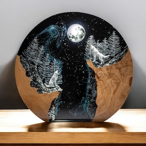 Puede incluir: Una obra de arte redonda de madera que representa una escena nocturna con dos lobos blancos aullando a la luna llena. El fondo es un cielo estrellado azul oscuro y negro con &aacute;rboles blancos y un r&iacute;o. La base es de madera marr&oacute;n claro.
