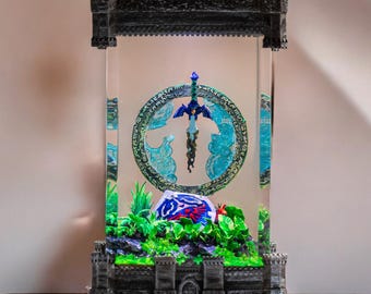 Puede incluir: Una vitrina de vidrio con una base de piedra gris y una espada azul y plateada en su interior. La espada está suspendida en un círculo con un diseño azul y verde. Hay una pequeña planta verde y una roca roja y azul en la base de la vitrina.