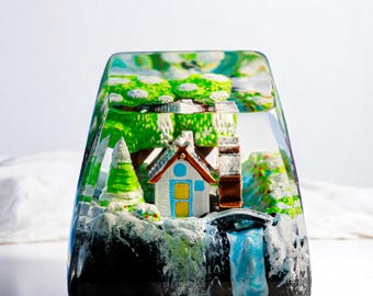 Puede incluir: Una pieza de arte de resina transparente de forma rectangular, que representa un paisaje en miniatura. La escena incluye una pequeña casa con una puerta azul, una cascada, árboles y un puente. La obra de arte está encapsulada en resina transparente y brillante.