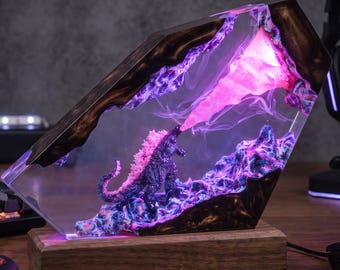 May include: A resin and wood lamp featuring a miniature Godzilla figure emitting a bright pink light beam. The scene is encased in a clear, angular resin block with dark wood accents, set upon a rectangular wooden base that illuminates the display.