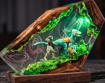 May include: A decorative lamp featuring a natural wood frame encasing a clear resin block. Inside the resin, a miniature human figure stands beside a blue dinosaur with a saddle, surrounded by green mossy textures and swirling white smoke effects.