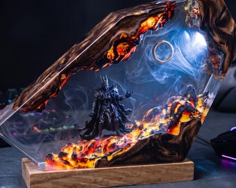 May include: A resin and wood display piece featuring a dark armored figure standing on a fiery, lava-like base. A golden ring is suspended in the upper section, surrounded by swirling white smoke and illuminated by internal blue and orange lighting.