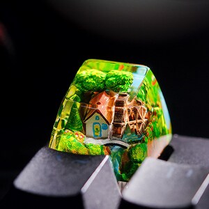 Könnte beinhalten: Eine farbenfrohe, individuelle Tastenkappe mit einer Miniatur-Dorfszene, die ein Haus, ein Wasserrad und üppiges Grün zeigt. Die transparente Tastenkappe befindet sich auf einer schwarzen Tastatur.