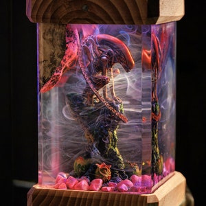 May include: A wooden lamp base with a clear glass cylinder containing a red and black alien figurine, surrounded by pink and white stones and white smoke.