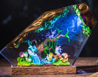 May include: A trapezoidal resin lamp featuring a miniature forest scene with multiple colorful creature figurines, including a central tree, green mossy base, and glowing blue internal lighting, all mounted on a rectangular wooden stand.