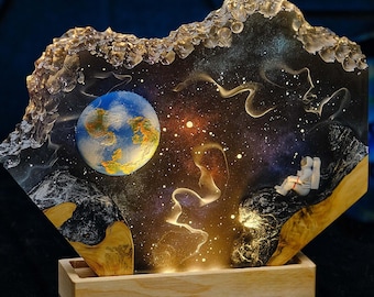 May include: A resin art piece depicting a galaxy scene with a blue planet, stars, and a white astronaut sitting on a rock. The piece is lit from behind, creating a glowing effect. The resin art is mounted on a wooden base.