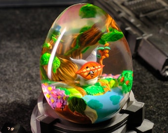 May include: A clear glass sphere with a miniature forest scene inside. The scene features a small, orange and white creature with a leafy green tree and flowers. The sphere is on a black base.
