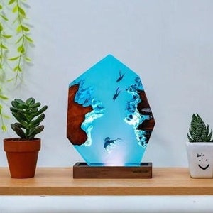 Könnte beinhalten: Eine blaue Harzlampe mit einem Holzsockel, die ein Design aus Wellen und Fischen zeigt. Die Lampe steht auf einem Holzregal mit zwei Topfpflanzen.