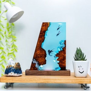 Könnte beinhalten: Dekorative Kunst aus Kunstharz mit einer blauen Ozeanszene mit Tauchern und einer braunen und weißen Küstenlinie. Das Kunstwerk steht auf einem Holzsockel. Eine kleine Sukkulente in einem weißen Topf und ein dekorativer Stein sind ebenfalls vorhanden.