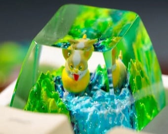 Puede incluir: Una tecla artesanal personalizada con una criatura amarilla y blanca en un paisaje en miniatura de colinas verdes y agua azul. La tecla está encapsulada en resina transparente con forma geométrica.