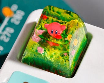 Puede incluir: Un tapón de tecla de resina verde y amarilla con un personaje rosa en su interior. El tapón de tecla está en un teclado blanco con teclas azules.