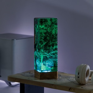 Könnte beinhalten: Eine grüne und blaue Harzlampe mit einem Holzsockel. Die Lampe ist eingeschaltet und strahlt ein warmes Licht aus.