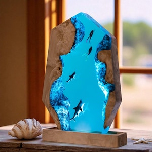 Puede incluir: Una escultura decorativa de madera con un centro de resina azul, que representa tiburones y buceadores. La escultura está sobre una base de madera y tiene una concha y una estrella de mar cerca. La madera tiene vetas naturales.