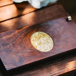 May include: A wooden box with a gold-colored, ornate design on the top. The box is made of dark wood and has a smooth, polished finish.