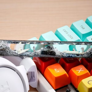 May include: A resin keyboard keycap featuring a black and white ocean scene with a sailboat and waves. The keycap is on top of a teal and white keyboard with orange and yellow keys.