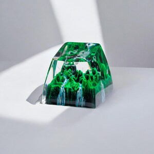 May include: A clear glass cube with a green and white landscape inside. The landscape features a waterfall and mountains.