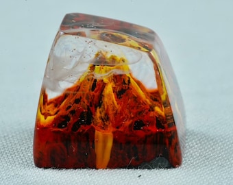 Op de afbeelding: Een transparante hars toetsenkap met een rood, oranje en geel vulkaanontwerp erin. De vulkaan heeft een witte, bewolkte top.