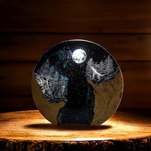 Puede incluir: Una obra de arte redonda de madera que representa una escena de bosque nocturno con dos lobos aullando a la luna llena. La obra tiene un fondo azul oscuro y negro con &aacute;rboles y estrellas blancas. Se exhibe sobre una superficie de madera.