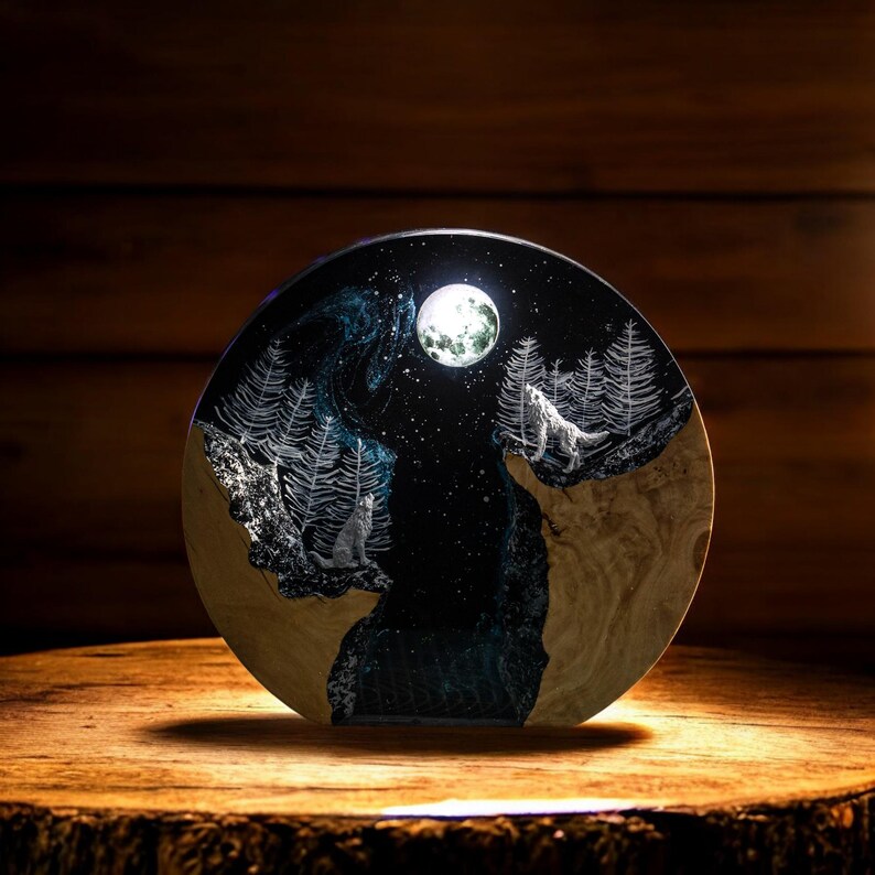 Puede incluir: Una obra de arte redonda de madera que representa una escena de bosque nocturno con dos lobos aullando a la luna llena. La obra tiene un fondo azul oscuro y negro con &aacute;rboles y estrellas blancas. Se exhibe sobre una superficie de madera.