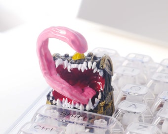 May include: A small, gold-colored treasure chest with a pink tongue sticking out of the open mouth. The chest is sitting on a clear keyboard with keys labeled "Ctrl", "Shift", "Win", and "Alt".
