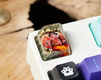 May include: A clear resin keycap with a red dragon figurine inside. The dragon is surrounded by orange, yellow, and green resin. The keycap is sitting on a white keyboard.