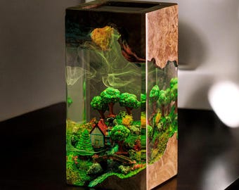 Puede incluir: Una pieza de arte rectangular de resina que presenta un paisaje en miniatura con una casa, árboles y un puente. Los lados son veteados de madera, y el interior tiene una combinación de colores verde y amarillo, creando un efecto de diorama. La pieza está sobre una superficie oscura.