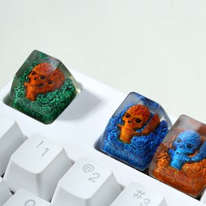 May include: Three clear resin keycaps with orange and blue skulls surrounded by flowers. The keycaps are on a white keyboard.
