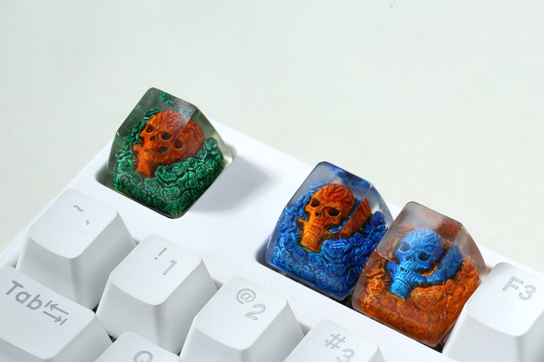 May include: Three clear resin keycaps with orange and blue skulls surrounded by flowers. The keycaps are on a white keyboard.