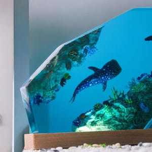 May include: A blue resin sculpture of a whale shark swimming in a blue ocean with coral reefs and other sea life. The sculpture is encased in clear resin and mounted on a wooden base.