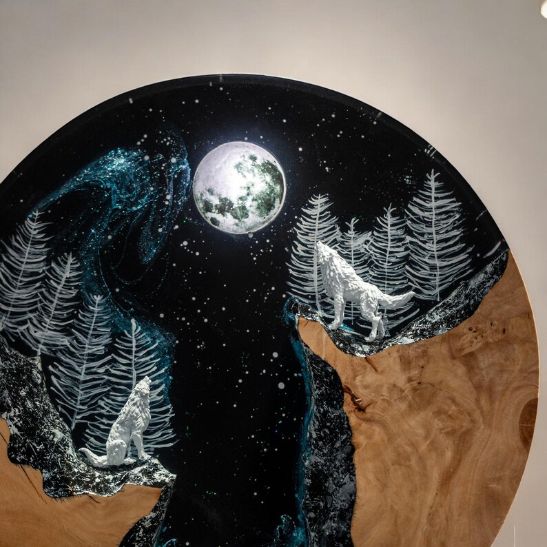 Puede incluir: Obra de arte redonda de madera que representa una escena nocturna con dos lobos blancos aullando a la luna llena. El fondo es azul oscuro con estrellas blancas y &aacute;rboles blancos estilizados. La parte inferior tiene un patr&oacute;n de vetas de madera.